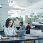 Stampante multifunzione a colori Xerox® C325 in una situazione d'ufficio Donna professionista che digita sul laptop alla scrivania in ufficio accanto a una stampante Xerox® C325